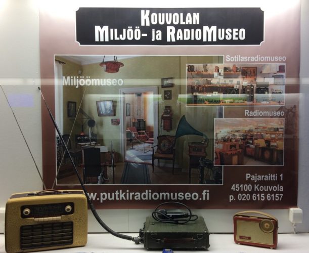 Display case for the Miljöö museum and radio museum in Kouvola railway station, Finland. Three radios displayed in front of a poster for the museums.